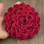 A hand holding a green crochet rose coaster, with white daisies in the background."