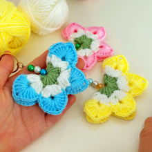 A handmade crochet flower keychain. The flower is composed of various pastel colors, including pink, blue, and green, giving it a vibrant appearance. The petals are textured, showcasing a crochet design that enhances its visual appeal.