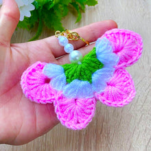 A handmade crochet flower keychain. The flower is composed of various pastel colors, including pink, blue, and green, giving it a vibrant appearance. The petals are textured, showcasing a crochet design that enhances its visual appeal.