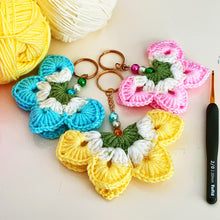 A handmade crochet flower keychain. The flower is composed of various pastel colors, including pink, blue, and green, giving it a vibrant appearance. The petals are textured, showcasing a crochet design that enhances its visual appeal.