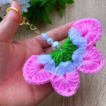 A handmade crochet flower keychain. The flower is composed of various pastel colors, including pink, blue, and green, giving it a vibrant appearance. The petals are textured, showcasing a crochet design that enhances its visual appeal.