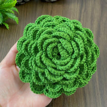 A hand holding a green crochet rose coaster, with white daisies in the background."
