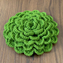 A hand holding a green crochet rose coaster, with white daisies in the background."