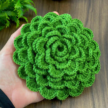 A hand holding a green crochet rose coaster, with white daisies in the background."