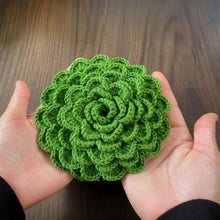 A hand holding a green crochet rose coaster, with white daisies in the background."